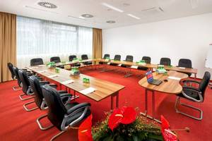 Konferenzraum im Star Inn Hotel Wien Schönbrunn mit langen Tischen, schwarzen Stühlen und roten Teppich.
