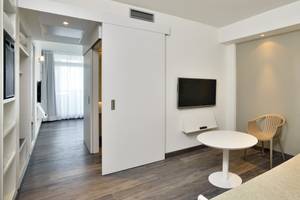 Innenansicht eines Zimmers im Sol Principe Hotel mit offenem Schiebetür, Flachbildfernseher an der Wand, weißem Tisch, Stuhl und Stehlampe.
