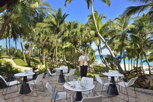Außenbereich des Melia Las Americas Hotels mit runden Tischen, Stühlen, und einem Kellner, umgeben von Palmen und Meerblick.