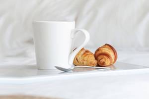 Ein weißer Kaffeebecher, zwei Croissants und ein Löffel auf einem Tablett auf einem Bett.