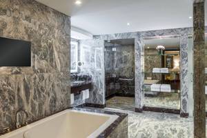 Badezimmer des Sheraton Oman Hotels mit Marmorwänden, Badewanne, Glasdusche und Fernseher.
