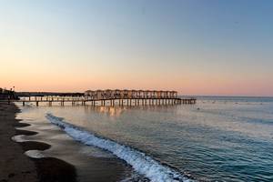 Strand mit Pier bei Sonnenuntergang, Wasser und Wellen, Gebäude im Hintergrund