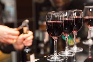Mehrere Gläser Rotwein stehen auf einer Bar, während jemand im Hintergrund eine Flasche öffnet.