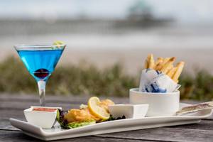 Cocktail, Fisch und Chips auf einem weißen Teller am Strand.