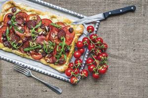 Eine Pizze mit Tomaten und Rucola auf einem Tablett neben Tomaten und einem Messer