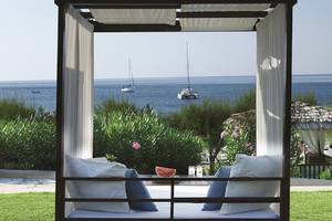 Ein Bett im Freien mit Blick auf das Meer und Segelboote, umgeben von Pflanzen und einem kleinen Tisch mit einer Wassermelone.