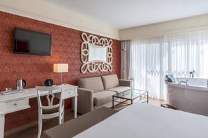 Luxuriöses Zimmer im Barcelo Corralejo Bay Hotel mit rotem Wanddesign, Fernseher, Schreibtisch, Couch, Badewanne und großer Fenster mit Vorhängen.