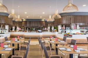 Innenansicht des Restaurants im Barcelo Corralejo Bay Hotel mit geometrischen Pendelleuchten, hölzernen Elementen und gedeckten Tischen.