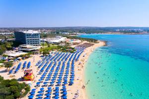 Luftaufnahme des Asterias Beach Hotels mit vielen Sonnenschirmen und Gästen am Strand.