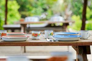 Ein Holztisch mit Geschirr und Besteck auf der Terrasse, hinter dem ein Garten und Tische und Bänke sind