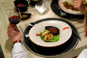 Ein Mann trinkt Rotwein in einem Glas während er ein Gericht mit Salat und Sauce genießt.