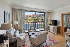 Innenansicht der Suite im Hilton Cabo Verde Sal Resort mit Wohnzimmer, Esstisch, Fernseher und Balkon.