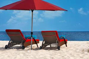 Zwei rote Sonnenliegen mit Kissen und einem roten Sonnenschirm auf dem Strand am Hilton Cabo Verde Sal Resort.