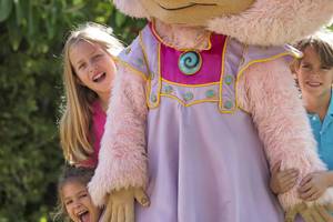 Drei Kinder, zwei Mädchen und ein Junge, posieren mit einem pinken Mädchen Maskottchen in einem Garten.