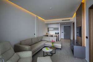 Wohnzimmer im Cavo Maris Beach Hotel mit L-förmiger Couch, Tisch, Fernseher und Küche im Hintergrund.