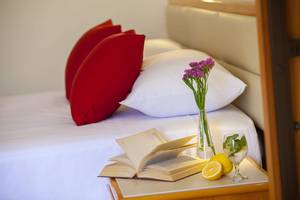 Ein Bett im Queens Bay Hotel mit roten Kissen, einer offenen Buche, einer Vase mit Blumen, einem Glas mit Limonade und Zitronen.