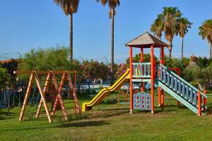 Spielplatz im Ilios K. Village Resort mit Rutsche, Klettergerüst und Pool im Hintergrund