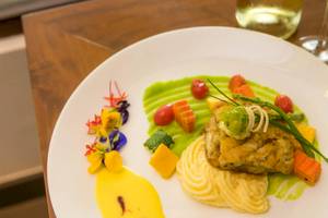 Ein Gericht auf einem weißen Teller, bestehend aus Fisch, Kartoffelpüree und Gemüse, mit einer grünen Sauce und dekorativen Blumen.