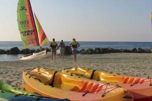 Segelboot am Strand von Baia del Sole mit drei Personen in Schwimmwesten und Kajaks auf dem Sand.