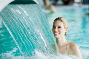 Frau im Schwimmbad unter einem Wasserfall, lächelt ins Bild