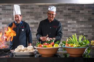 Zwei Küchenchefs des Alexander House stehen in der Küche und bereiten Essen zu, mit einem Koch, der Feuer benutzt, und Gemüse und Zutaten auf dem Tisch.