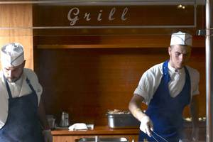 Zwei Köche im Grillbereich des Serita Beach Hotels, einer in Uniform, der andere in einer Haube, beide konzentrieren sich auf die Zubereitung von Speisen.