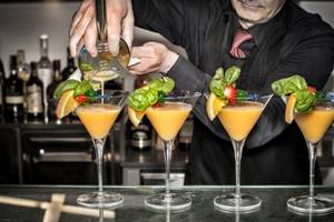 Ein Barkeeper serviert Cocktails auf einer Bar, vier Gläser mit verschiedenen Früchten und Kräutern.