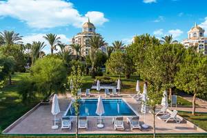 Ansicht des Delphin BE Grand Resort mit Pool und Palmen unter blauem Himmel. Entspannte Atmosphäre im Garten.