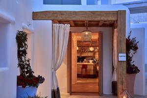 Eingang zum Mykonos Ammos Hotel mit hölzernem Vordach, offenen Türen und Innenansicht des Barsbereichs mit roten Stühlen und Blumentöpfen.