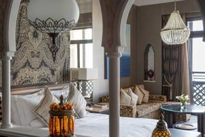 Innenansicht des Schlafzimmers im Jumeirah Mina A'Salam Hotel mit großen Bögen, Kronleuchtern, einem Bett, einem Tisch und einem Sofa.