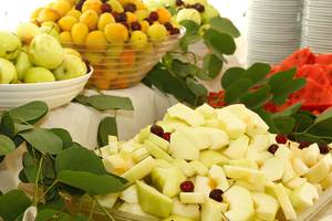 Ein Obstbuffet auf einem Tisch mit verschiedenen Früchten wie Trauben, Äpfeln, Aprikosen und Melonen, dekoriert mit grünen Blättern.