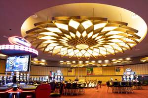 Innenbereich eines Casinos mit Spieltischen, Spielautomaten und einer großen, blumigen Lampe an der Decke.