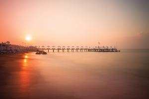 Ein Strand mit einem Pier und dem Sonnenuntergang im Hintergrund, reflektiert auf dem Wasser.