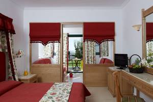 Innenansicht eines Zimmers im Mitsis Rinela Beach Hotel mit roten Vorhängen, einem Bett und einem Schreibtisch.