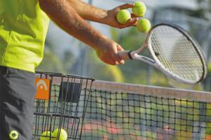 Tennisspieler hält Tennisschläger und Tennisbälle auf einem Tennisplatz mit Netz im Hintergrund.