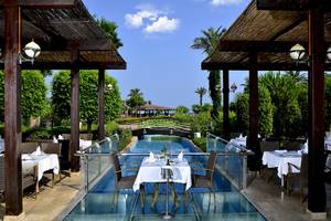 Pool-Terrasse im Miracle Resort mit Tisch und Stühlen, umgeben von Pflanzen und einer Brücke.
