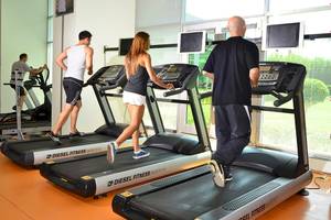 Fitnessstudio mit mehreren Personen, die auf Laufbändern trainieren, darunter ein Mann in einem weißen Tanktop.