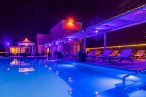 Beleuchteter Poolbereich des AX The Palace Hotels bei Nacht mit Lounge-Stühlen und bunten Lichtern.