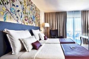 Doppelbett-Zimmer im AX The Palace Hotel mit kunstvollen Wandgemälden, zwei Betten, Tisch, Lampen und Stühlen.
