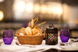 Ein Korb mit Brot und Snacks steht auf einem Tisch mit zwei violetten Glaesern und Besteck.