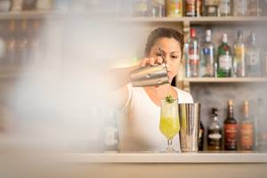 Frau mischt einen Cocktail an einer Bar mit vielen Flaschen im Hintergrund.