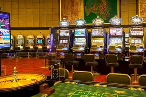 Innenbereich eines Casinos mit Spielautomaten, Roulettetisch und Stühlen.