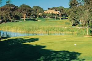 Golfplatz des Cornelia Diamond Golf Resort & Spa mit einem Gebäude im Hintergrund und einem See vor der Anlage.
