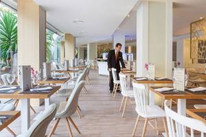 Innenansicht des Restaurants im Hotel Tigotan Lovers & Friends Playa de las Americas mit gedeckten Tischen und einem Kellner, der sich um einen Gast kümmert.