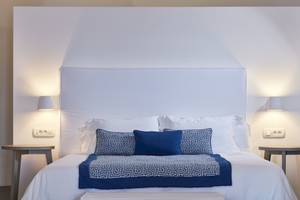 Bett im Katikies Chromata Santorini Hotelzimmer mit weißen Wänden, blauem und weißem Bettzeug und Nachttischen.