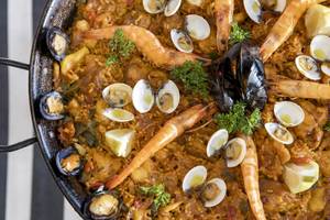 Ein großer Topf Paella mit Garnelen, Muscheln und Reis auf einem schwarzen und weißen gestreiften Hintergrund.