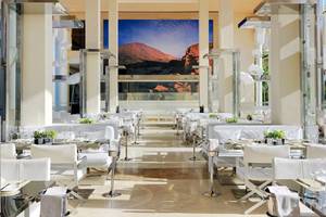 Innenbereich des Gran Melia Palacio de Isora Restaurants mit vielen weißen Tischen und Stühlen und einer großen Glaswand.