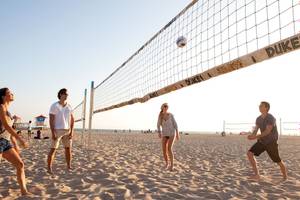 Vier Personen spielen Volleyball am Strand mit einem Netz, das Duke und Sandy & Duke beschriftet ist.