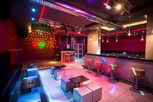 Innenansicht einer Disco mit Bar, Barhockern und Tischchen sowie farbiger Beleuchtung.