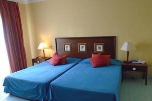 Doppelbett-Zimmer im ROC Arenas Doradas Hotel mit gelben Wänden, blauen Laken und roten Kissen.